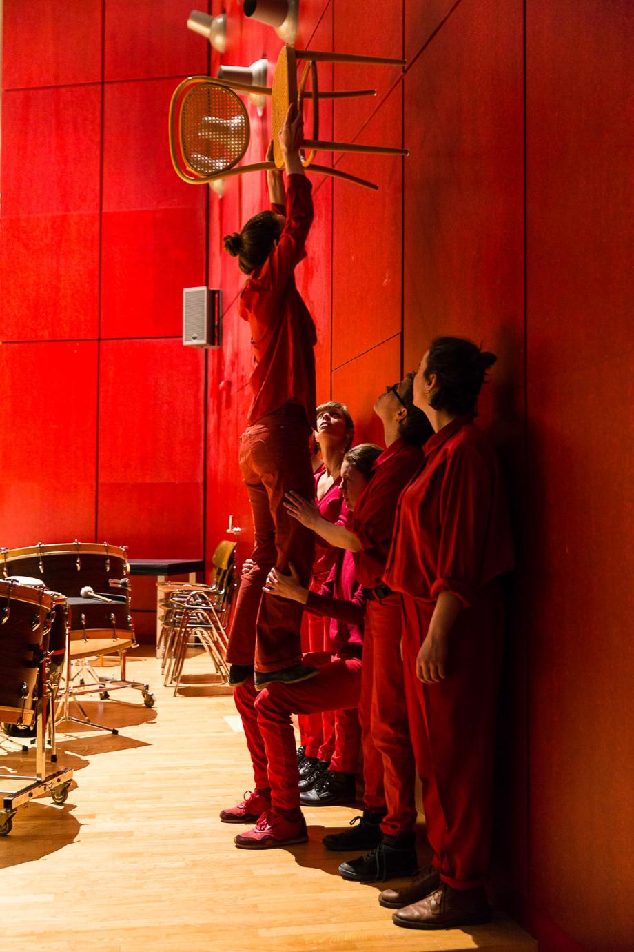 Studierende des Figurentheaters, alle in rot gekleidet, stehen an der Konzertsaalwand und versuchen einen Stuhl in die Luft zu heben.