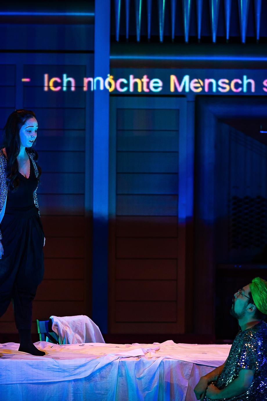 Ein Pärchen schaut sich aus der Distanz an. Im Hintergrund ist auf dem Prospekt zu lesen "Ich möchte Mensch sein". Die Szene ist atmosphärisch dunkel gehalten.