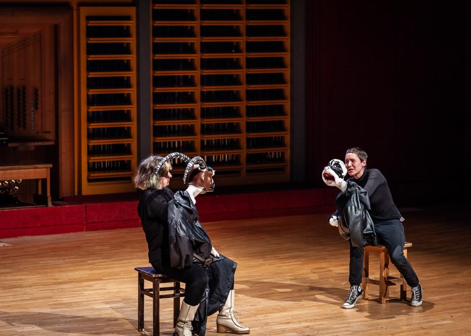 Zwei Studierende, die auf schwarzen Schmeln sitzend und mit jeweils einer Handmaske ausgestattet sind, auf dem Podium des Konzertsaals. 