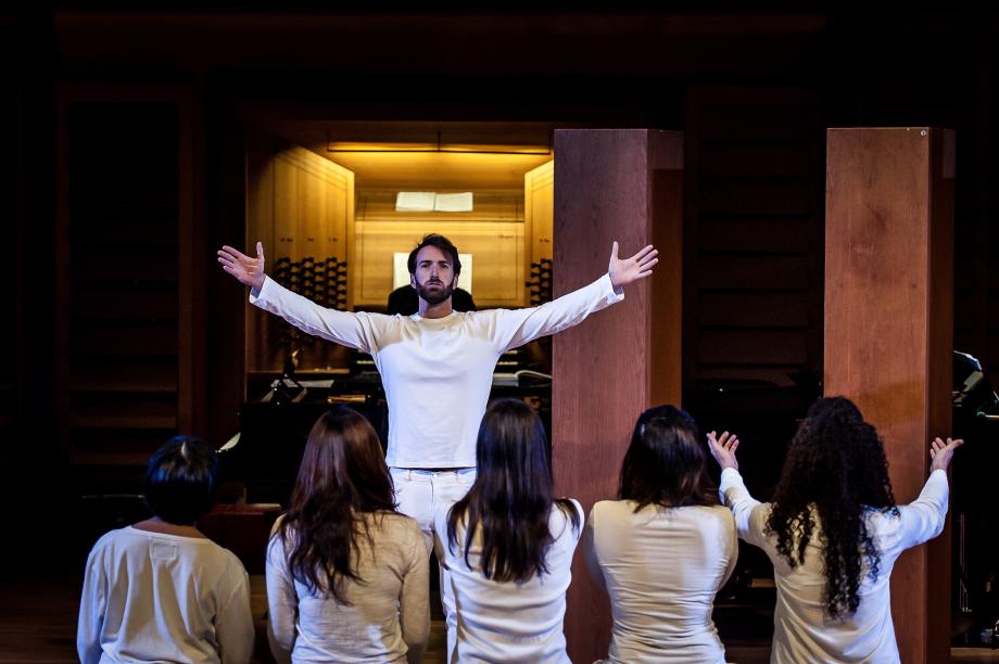 Opernszne mit einem Studierenden, der beide Arme weit ausbreitet, beobachtet von fünf Frauen, die vor ihm knien und alle in weiß gekleidet sind.