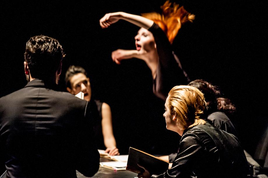 Szenisch agierende Studierende, die mit Büchern um einen Tisch sitzen, den Blickkontakt suchen. Im Hintergrund erhebt sich eine Studentin mit ausladener Armbewegung.