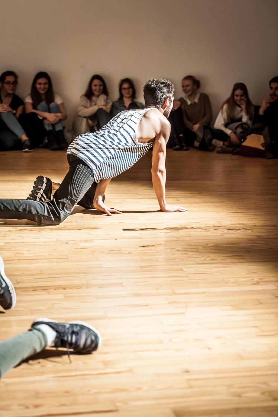 Auf allen Vieren agierender Student, der von hinten fotografiert wird. Man sieht das beobachtende, am Fußboden sitzende Publikum.