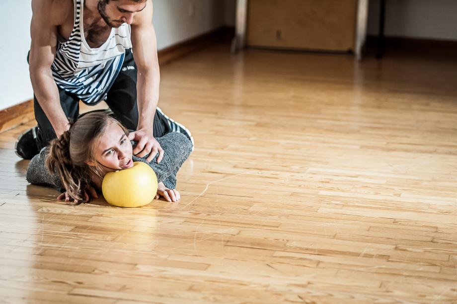 Schauspielszene, wo ein knieender Student eine Studentin auf einen gelben Ball drückt.