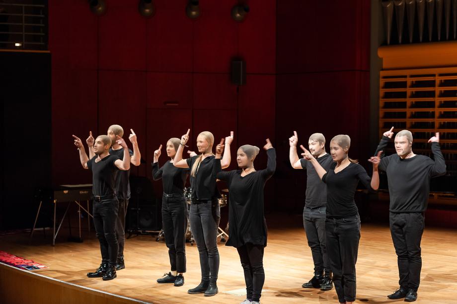 Sprechchor auf der Bühne des Konzertsaales. Alle Gesichter sind mit einer Strumpfhose überzogen. Der Chor agiert mit Hand- und Armbewegungen. Alle Mitwirkenden sind in schwarz gekleidet.