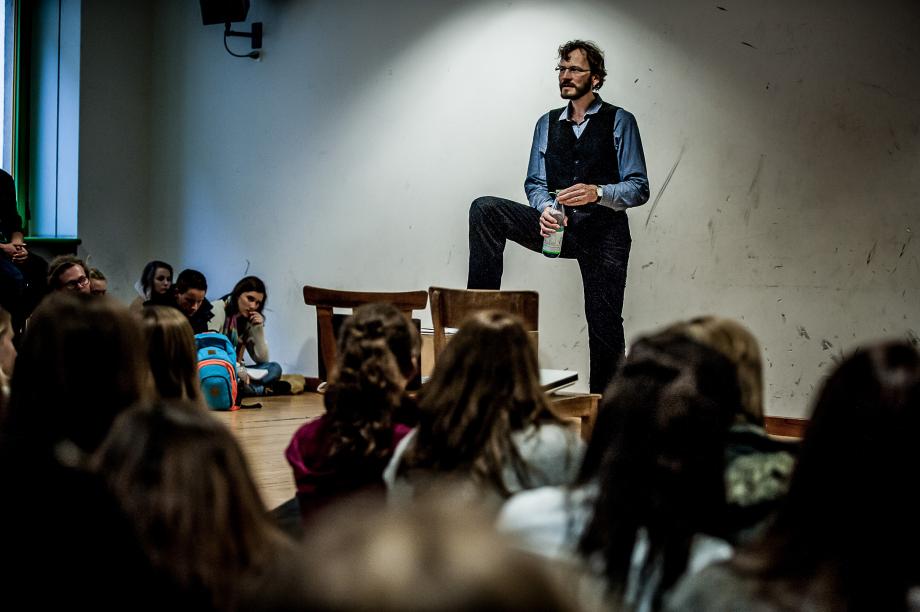 Schauspieldozent Frederik Zeugke bei einer Einführungsveranstaltung. Sein rechtes Bein ist auf eine Kiste gestützt. In der Hand hält er eine Wasserflasche und sucht dabei den Blickkontakt zum Plenum.