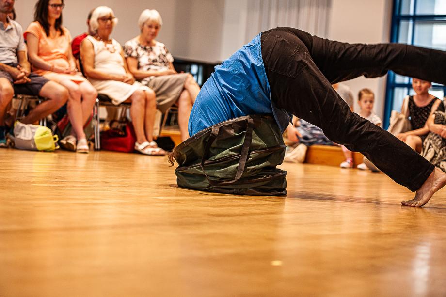 Ein Studierender des Figurentheater lehnt sich im Rahmen einer Performance in einen grünen Laubsack. Das Gesicht ist verdeckt, ausschließlich das leucht blaue Hemd und die teilweise abgewinkelten Beine sind zu sehen. Das Publikum beobachtet diese Szene.