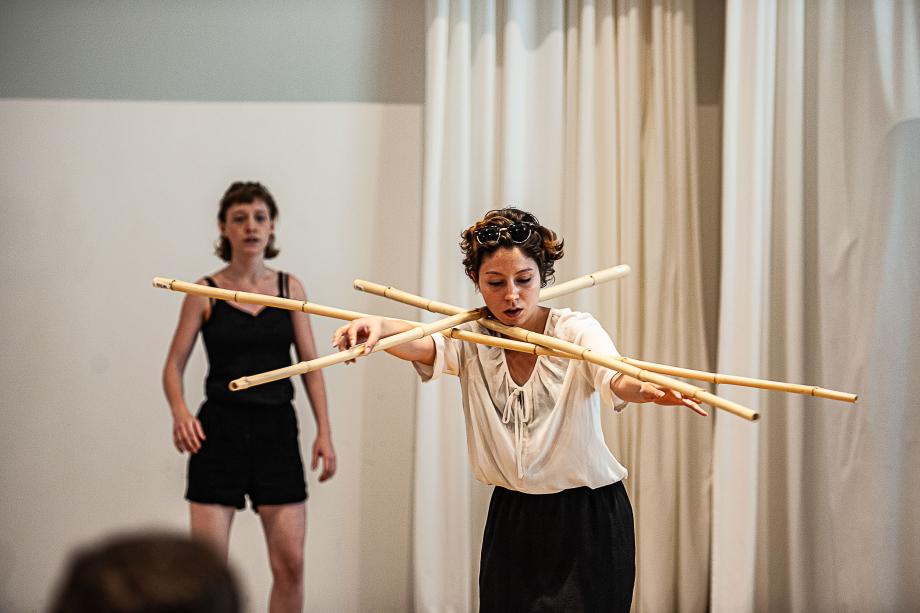 Balancearbeit einer Studierenden mit drei langen Bambusstäben, die üner ihre ausgestreckten Arme liegen. Im Hintergrund beobachtet eine Studierende diese Situation.