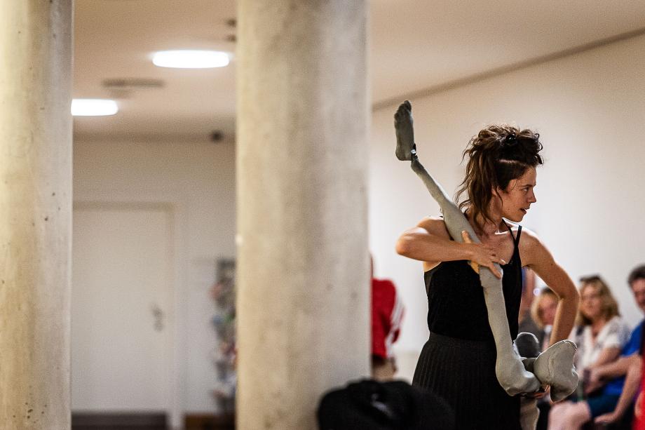 Studierende, die ausschließlich mit Beinen einer großen Puppe tanzt. Im Hintergrund sind Zuschauerrinnen und Zuschaue zu sehen, die diese Szene verfolgen.