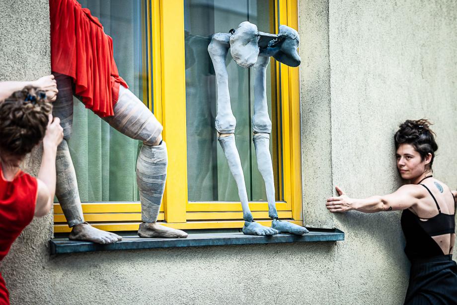 Im Fokus sind zwei Beingestelle von Puppen auf einer Fensterbank zu sehen. Von links und rechts nähern sich zwei Studierende, die sich eng an die Fassade drücken.