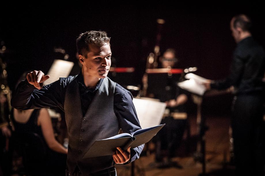 Student der Sprechkunst hält eine Mappe vor sich, den Blick in den Saal gerichtet. Im Hintergrund ist der Bühnenaufbau für ein Instrumentalensemble zu sehen.