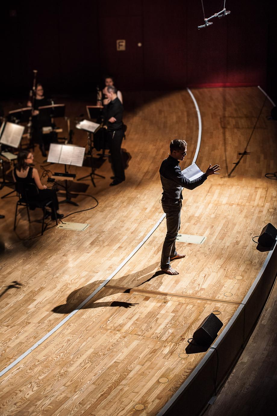 Sprecher mit klarer Haltung undd Ausrichtung auf das Publikum. Im PRofil aufgenommen und von Mitgliedern des Ensembles beobachtet wird.