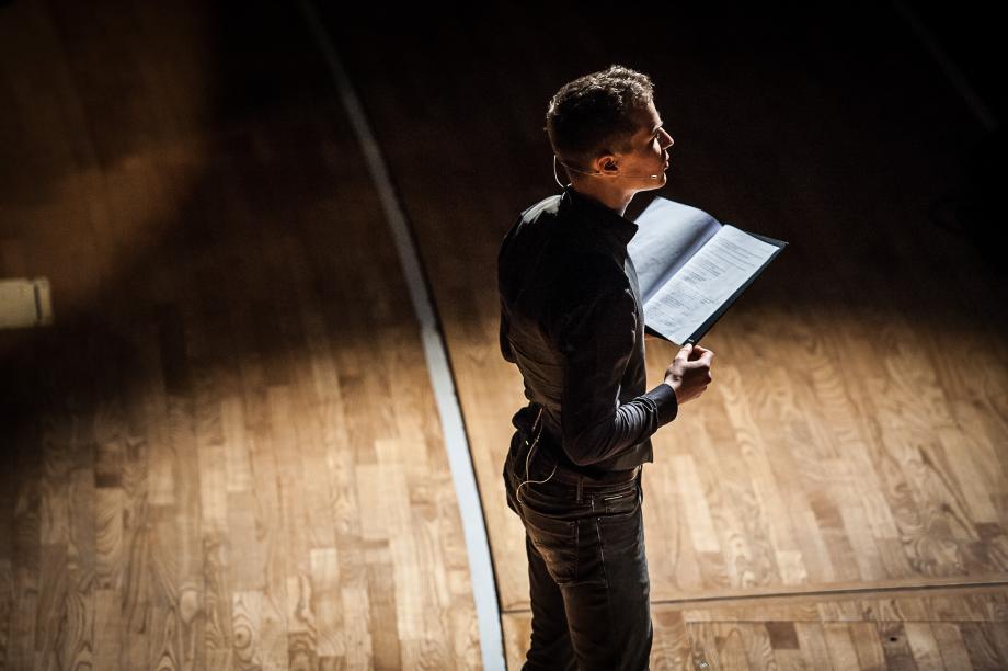 Detailaufnahme eines Sprechers, der im Lichtkegel auf der Bühne steht, mit einem Headset ausgestattet ist und eine Mappe in der Hand trägt.