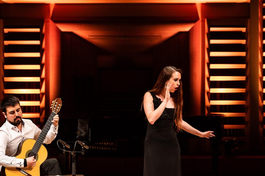 Gitarren und gesangsduo auf dem Podium. Die Sängerin im schwarzen Abendkleid dreht ihr Gesicht weg und flüstert imaginär einen dritten Person etwas zu.