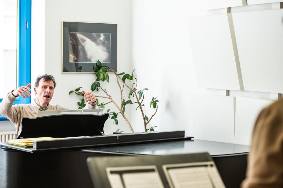 Gesangsdozent Thomas Scharr beim Unterrichten vom Flügel aus. Dabei hebt er beide Arme, um dirigierend die Gesangsphrase nachzubilden.