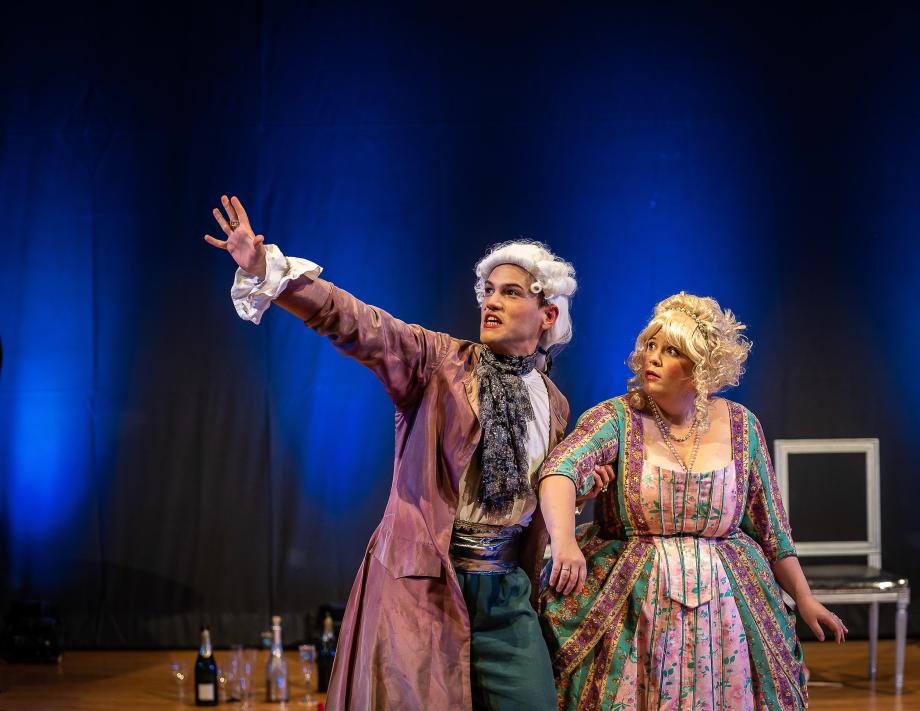 Zwei Studierende in Kostümen auf der Bühne. Sie schauen der Hand des Mannes nach, die nach oben zeigt.