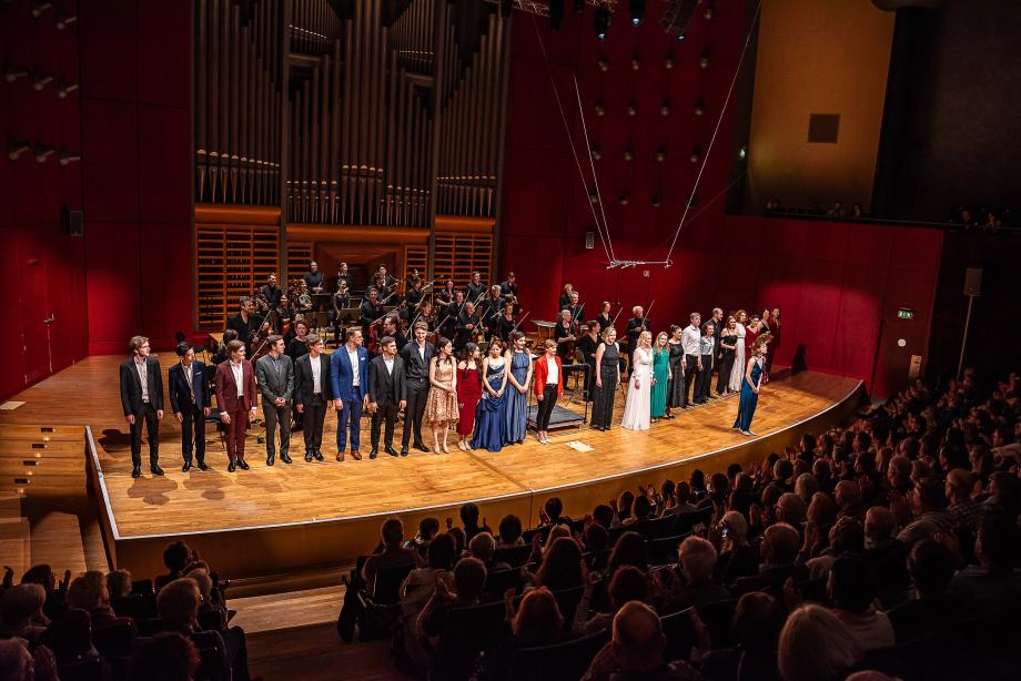 Großes Opernensemble beim Schlussapplaus mit Orchester. Im Vordergrund ist der gut besetzte Konzertsaal zu sehen.