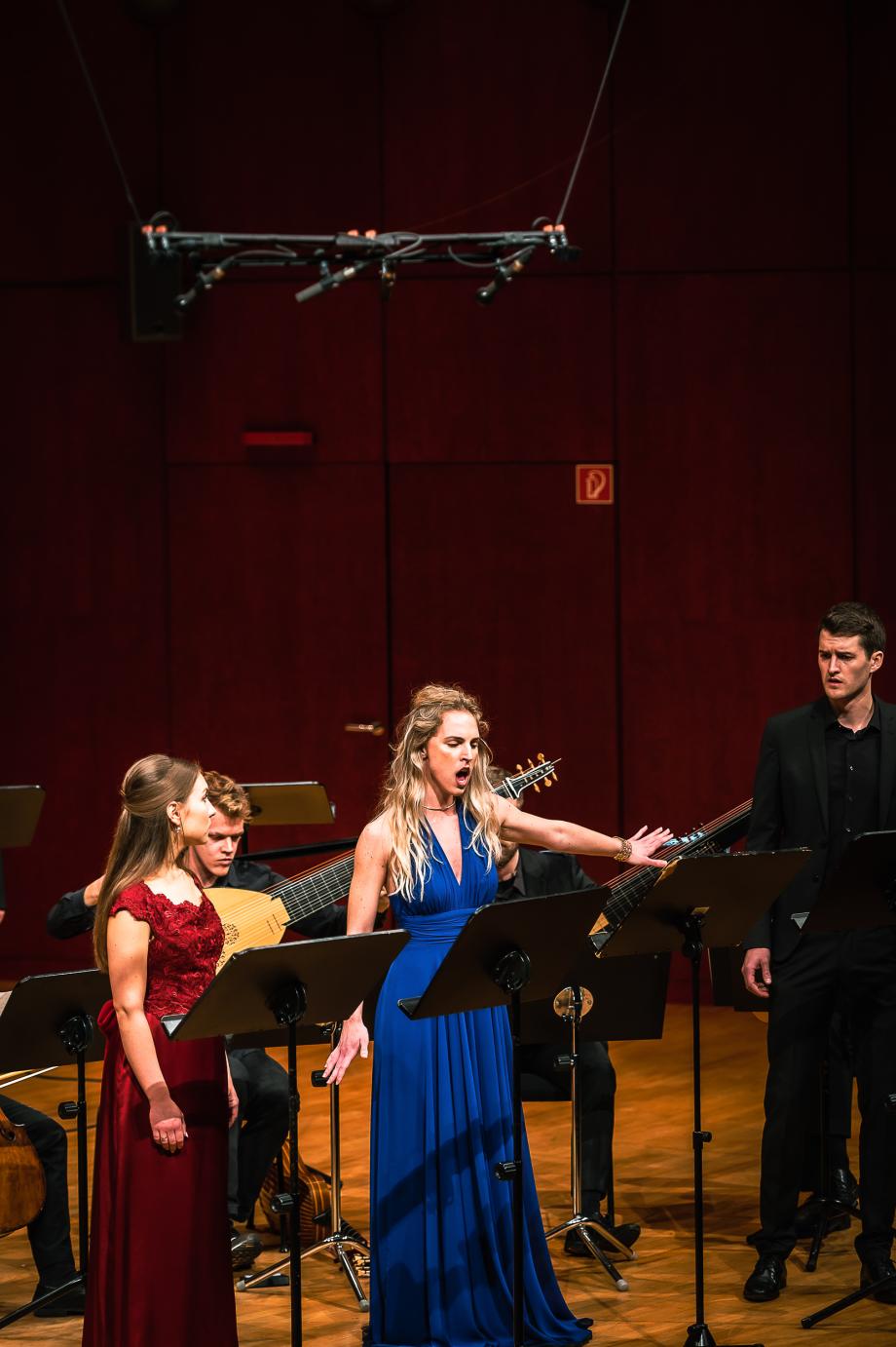 Sängerin im blauen Abendkleid mit großer, abwehrender Geste. Im Hintergrund musiziert ein Kammerensemble.