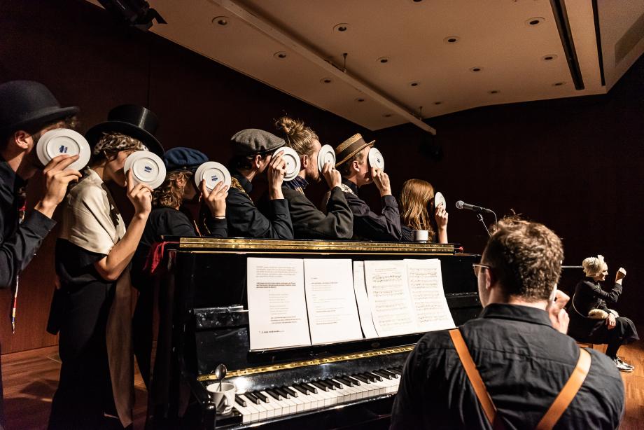Sieben Studierende, die sich mit ihren Ellenbogen am Resonanzkörper eines Klacieres abstützen. Ihre Gesichter sind mit Untertellern verdeckt. Der Pianist ist von hinten zu sehen.
