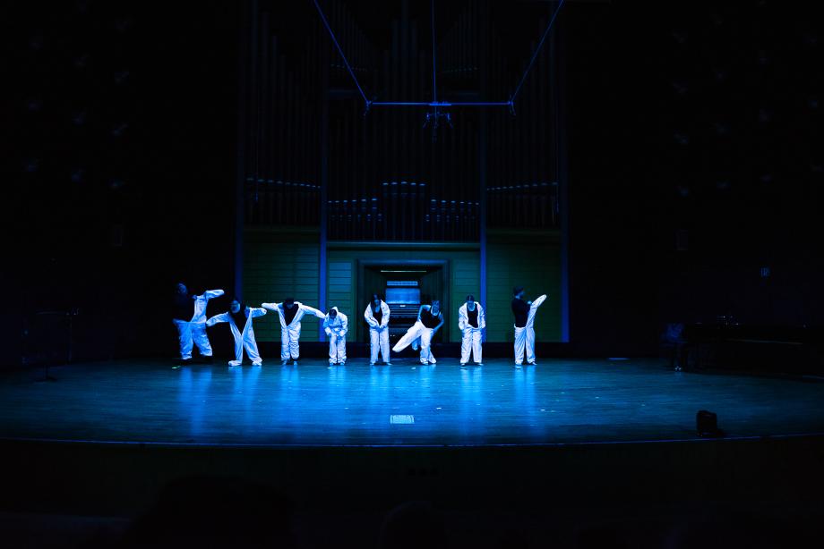 acht Studierende in weißen Maleranzügen. Die Bühne ist in blaues Licht getaucht.