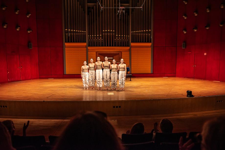 sechs Studentinnen in weißen Hosen und beigen Hemden stehen zum Applaus bereit.