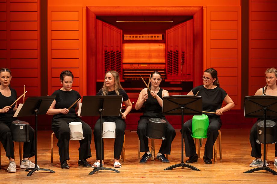 Studentinnen sitzen auf Stühlen vor Notenständern und spielen Schlagzeug auf Kunststoffeimern.