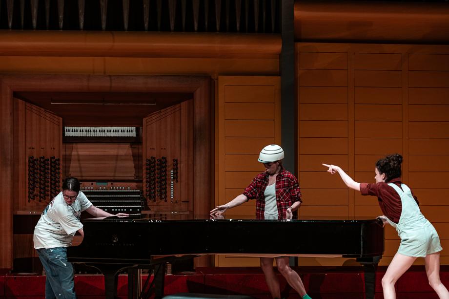 drei Studentinnen stehen an einem Flügel. Eine Studentin, die an der Seite und in der Mitte steht, trägt einen weßen Fahrradhelm