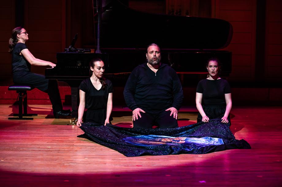 Drei studierende knien auf dem Boden vor einem glitzernden Tuch und singen. Hinter ihnen spielt eine Pianistin.