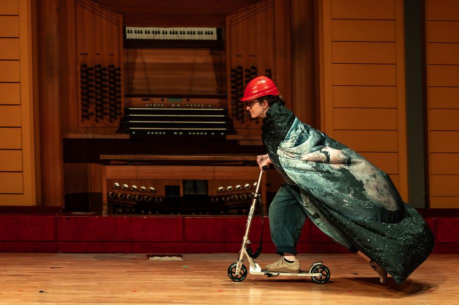 Studentin mit rotem Helm und Umhang rast auf einem Roller über die Bühne.