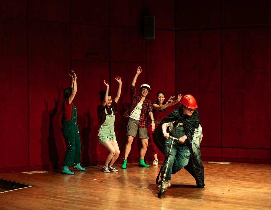 fünf Studentinnen auf der Bühne. Eine Studentin mit rotem Helm und einem Umhang fährt auf einem Roller. Die anderen Studentinnen jubeln ihr im Hintergrund zu.