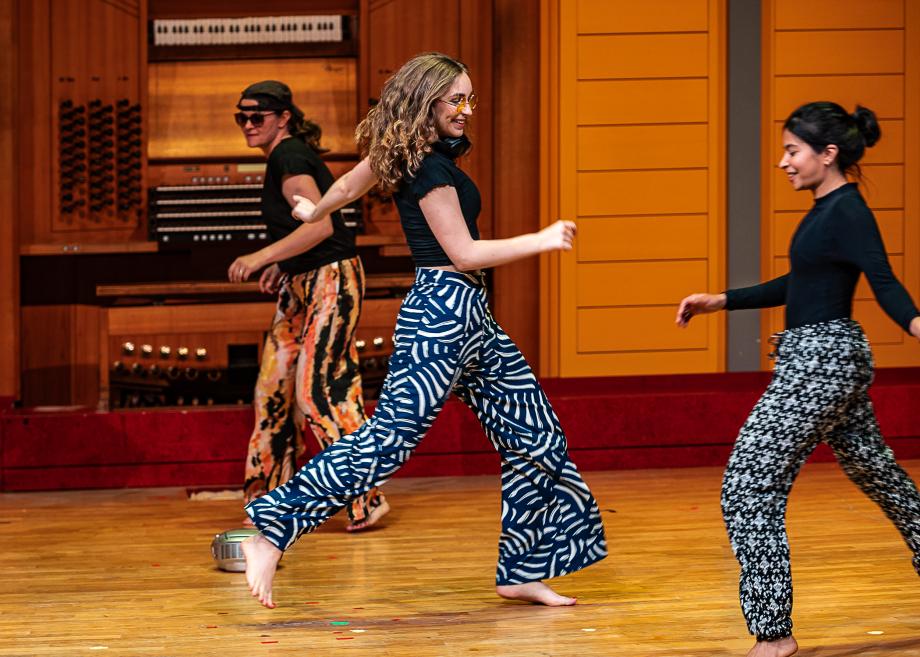 Drei Studentinnen in gemusterten Hosen tanzen barfuß über die Bühne.
