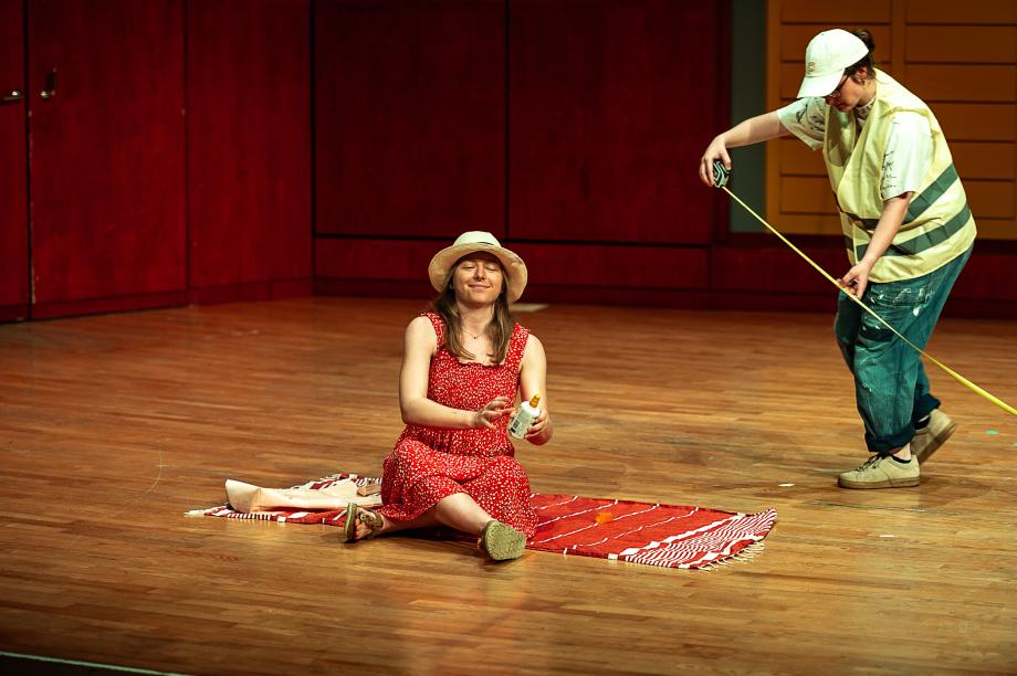 Studentin sitzt auf einem roten Strandhandtuch in einem roten Kleid mit Sonnenhut und cremt sich lächelnd ein. Hinter ihr misst eine Studentin, als Bauleitung verkleidet, mit einem Maßband etwas aus.