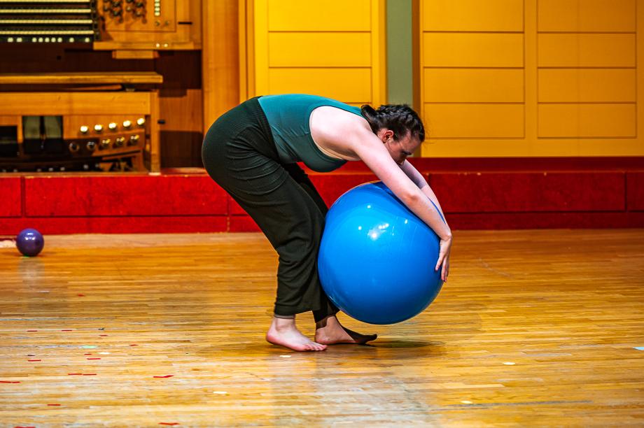 Eine barfüßige Studentin hält gebückt einen Gymnastikball an ihren Körper gepresst.