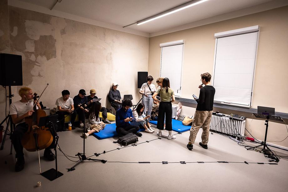 Studierende sitzen auf dem Boden in einem Seminarraum der Willy-Brandt-Straße. Zum teil sprechen sie miteinander, zum Teil liegt der Fokus auf dem Smartphone. Der Raum ist mit verschiedenen Kabeln verlegt und Performanceaktionen vorbereitet.