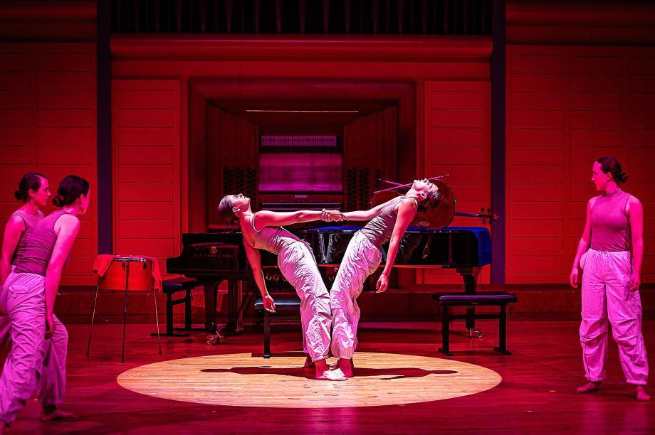 Im Lichtkgel stehen zwei Studierende, die sich an der rechten Hand festhalten und weitestgehend nach hinten fallen lassen. Die Szene ist im rötlichen Licht eingebunden. Das Duo wird von Studierenden beobachtet.