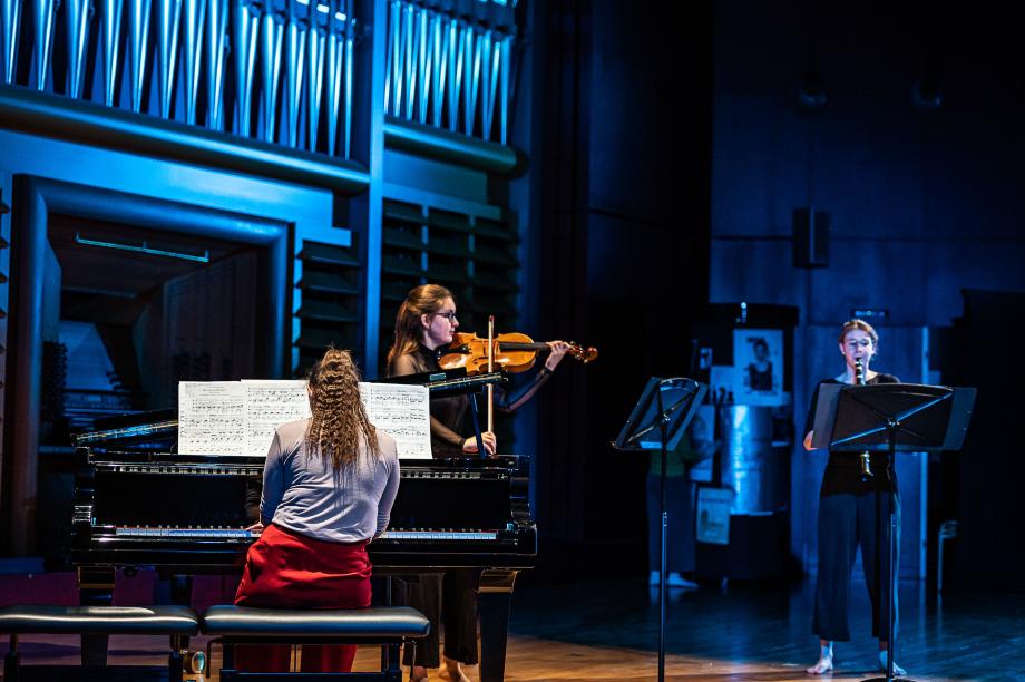 Kammermusiktrio auf dem Konzertsaalpodium. Szenisch dunkle Beleuchtung. Sowohl die Violinistin als auch die Klarinettistin musizieren barfuß.