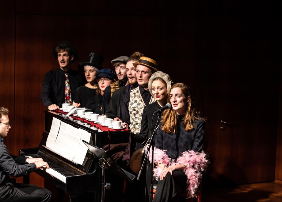 acht Menschen stehen vor Klavier und Pianisten aufgereiht. Eine Frau singt ins Mikrofon