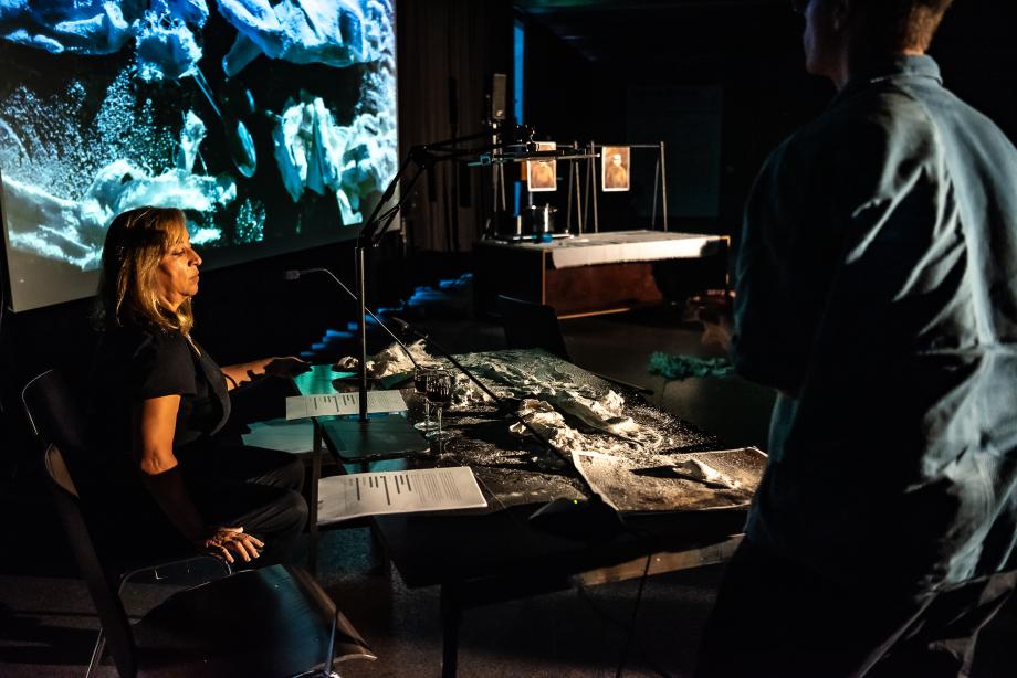 eine aufrecht sitzende Frau sitzt an einem Tisch vor einem Mikrophon. Vor ihr liegen zwei Texte. sie schaut nach unten. Im Hintergrund ist eine große Leinwand mit undefinierbaren Objekten drauf.