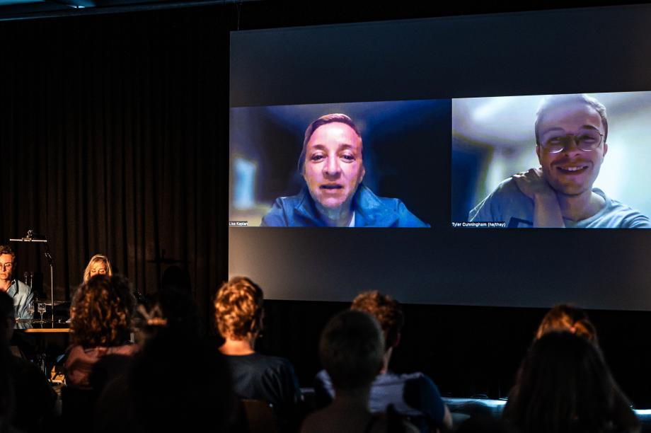 Publikum schaut auf eine Leinwand, auf der sich zwei Menschen in einem Videocall befinden.