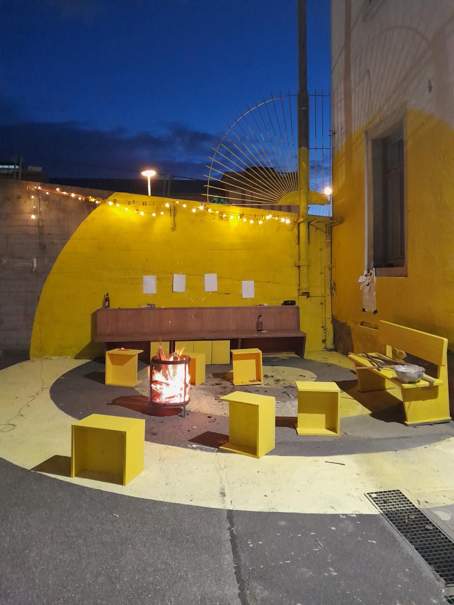 Außenbereich des Maker-Space mit selbstgebauten Sitzmöglichkeiten und Feuerstelle in der Abenddämmerung