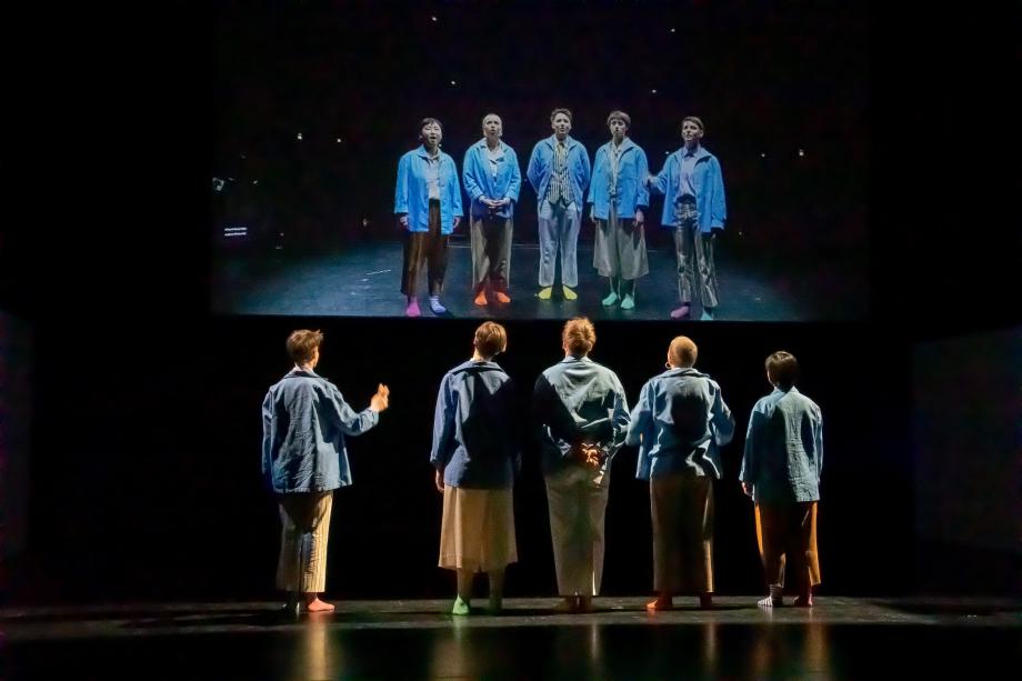 fünf Menschen, vom/von der Betrachter*in abgewendet, schauen sich selbst auf einem Bildschirm an.