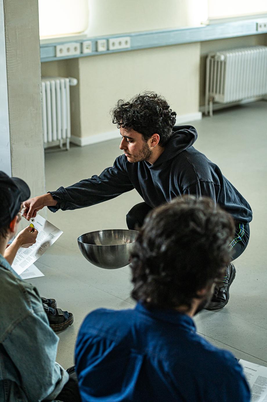 Mann hockt mit einer Schüssel in der Hand auf dem Boden und hält in der anderen Hand eine kleine Blume über einen Text, der in der Hand einer Frau ist.