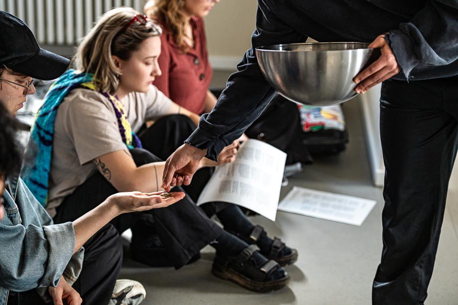 Mann mit Schüssel in der Hand gibt jemandem im Publikum, das auf dem Boden sitzt, etwas in die Hand.