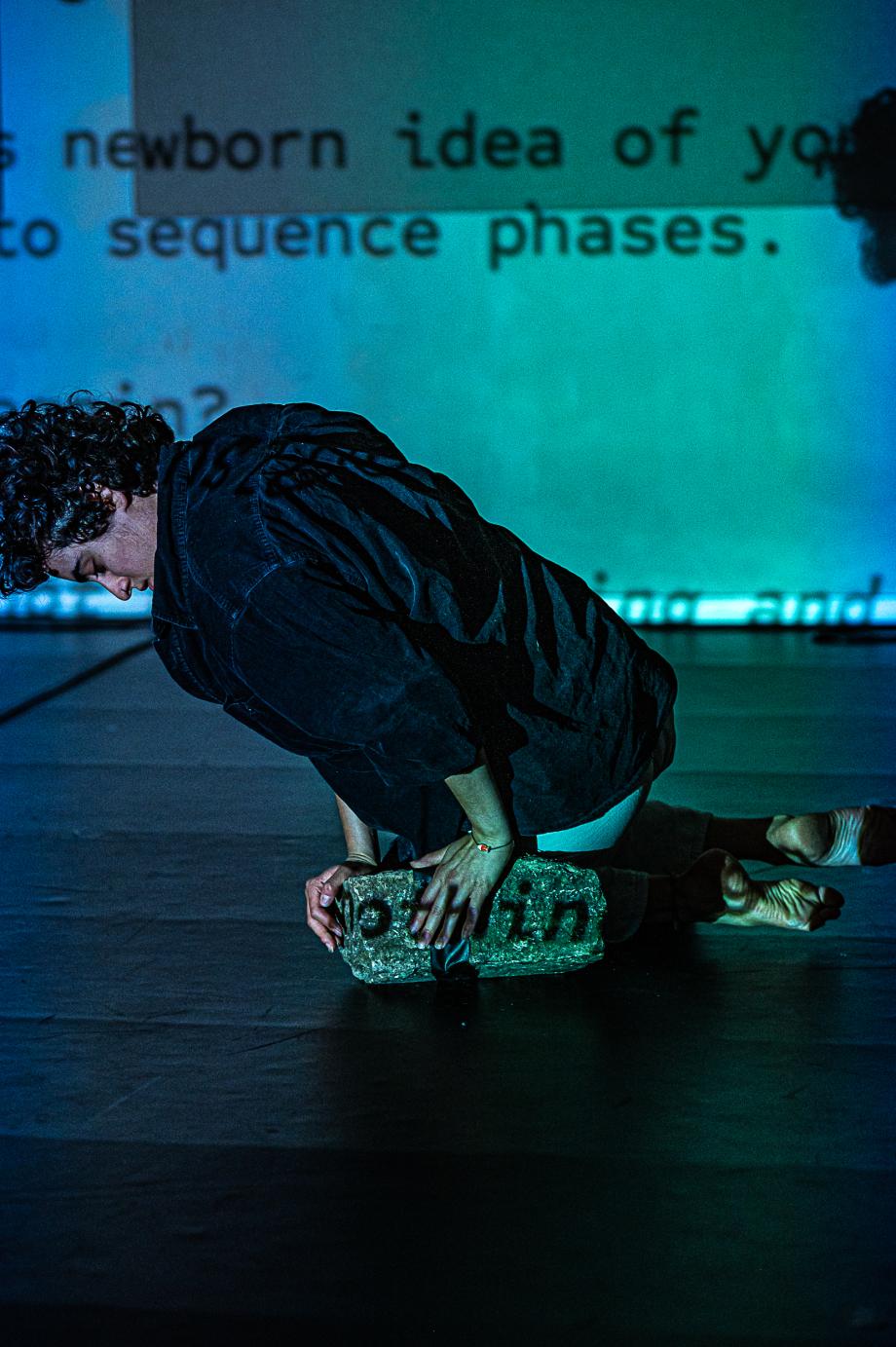 Frau vor Leinwand mit Text stützt sich auf einen Stein. Sie blickt nach unten.