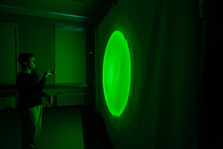 Mann projeziert grünen Kreis mit einer Taschenlampe auf eine Leinwand. Der Raum ist dunkel.