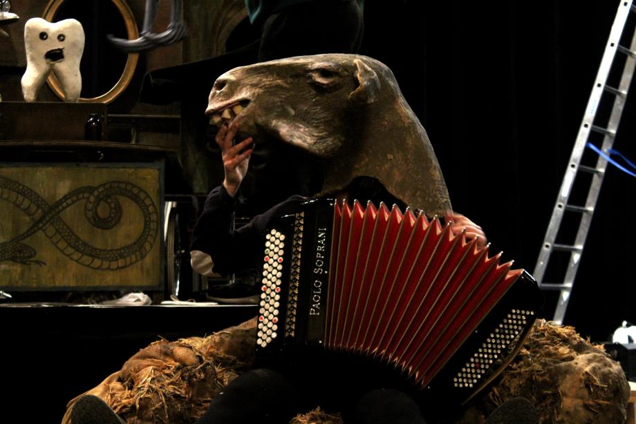 Ein Zahn mit einem Gesicht, der Munbd ist aufgerissen und ein Kopf eines Schafes und einer Ziehharmonika auf einem Strohberg.