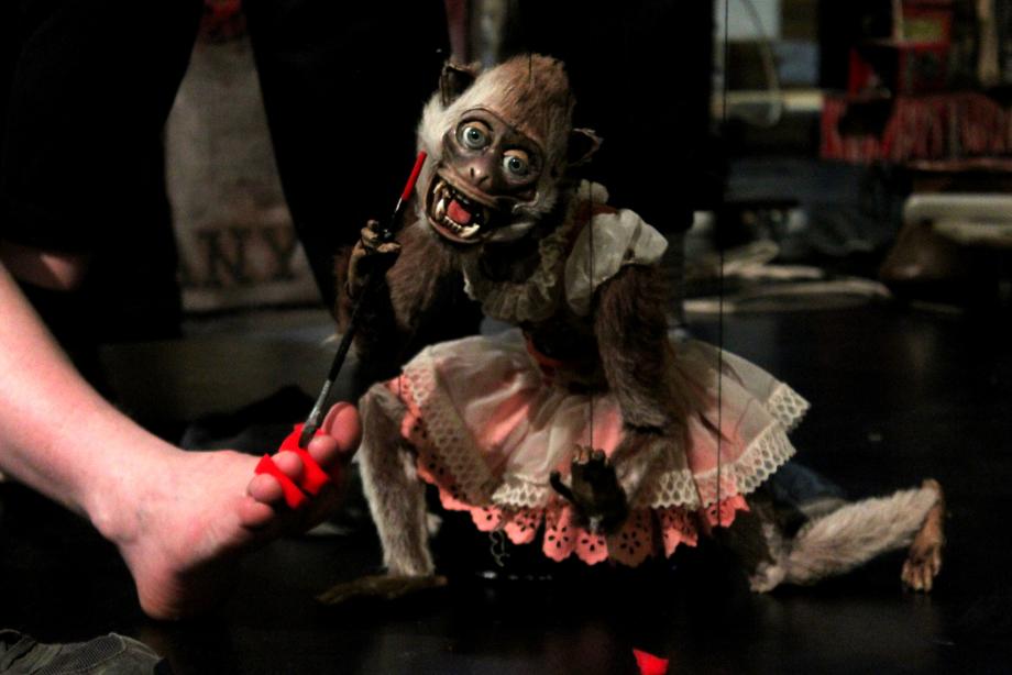 Affen-Marionette richtet Pinsel, den sie in der Pfote hält, auf einen nackten Fuß mit roten Schaumstoffteilchen zwischen den Zehen.