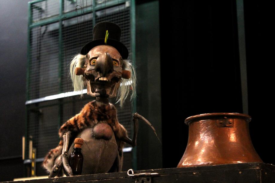 Marionette mit Flasche in der einen Hand, Sense in der anderen und Zylinder auf dem Kopf, reißt ihr Maul auf.