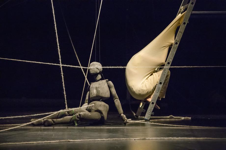 Figur mit Schnüren durch den Körper sitzt am Boden. Neben ihr rechts steht eine Leiter, auf der ein Mensch in einen beigen Stoff eingewickelt ist.