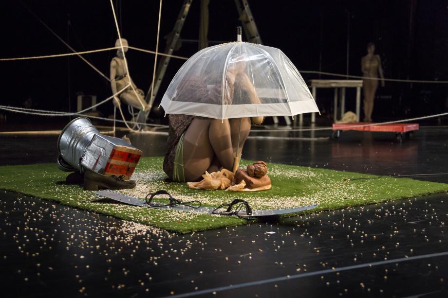 Frau sitzt auf grünem Kunstrasen, vor ihr ein Klumpen Ton. Sie sitzt zusammengekauert da und hält einen Regenschirm über sich. 
