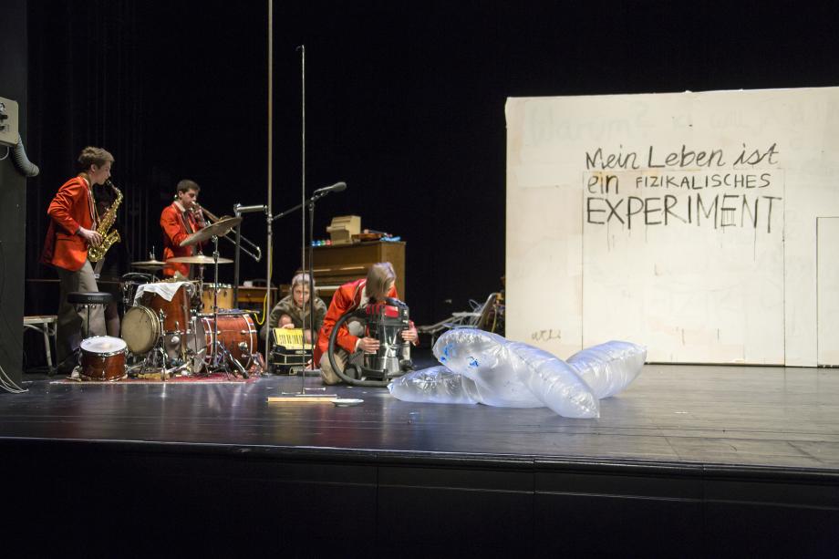 vier Menschen auf Bühne: drei in roten Jacken. Zwei davon spielen Instrumente, eine hält hockend ein Gerät, die andere Person hockt neben einem Instrument, trägt aber keine rote Jacke.